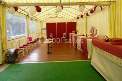 Tente avec table dressée pour un buffet, entourée de chaises blanches, installée sur une pelouse avec haies en arrière-plan.