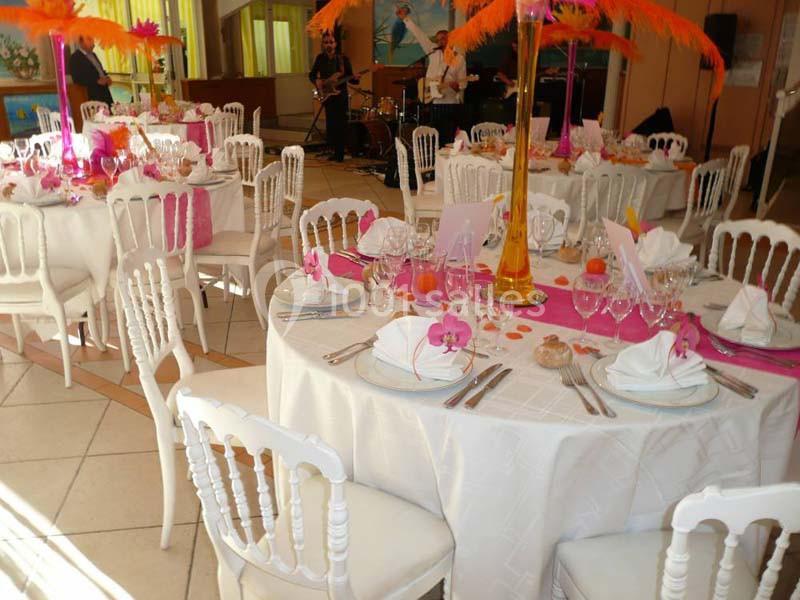 Salle décorée pour un événement avec tables dressées, nappes blanches, fleurs roses et plumes orange en centre de table.