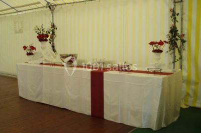 Tente avec table dressée pour un buffet, entourée de chaises blanches, installée sur une pelouse avec haies en arrière-plan.