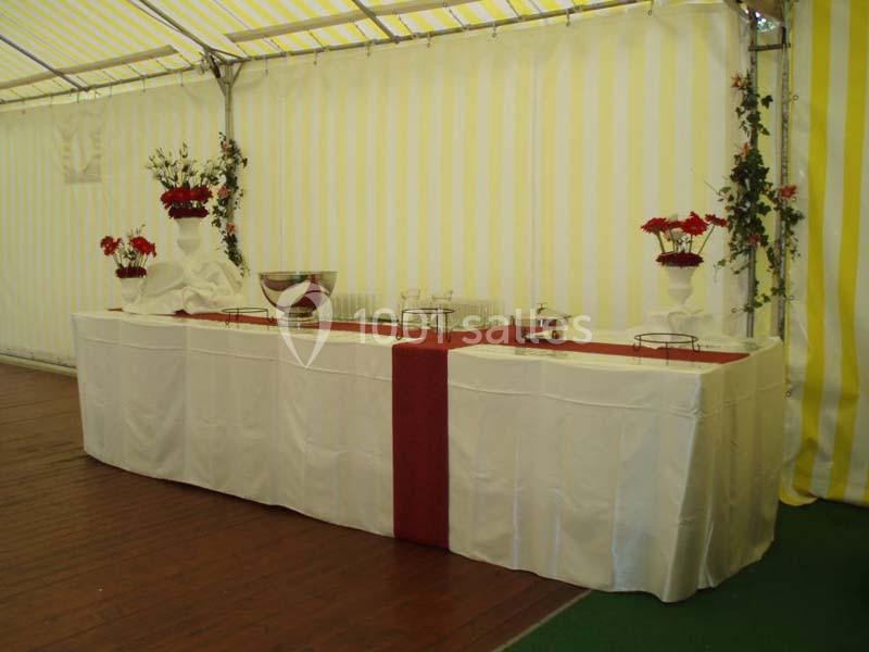 Table décorée avec nappes blanches, chemin de table rouge, vases de fleurs rouges et vaisselle, sous une tente rayée jaune…