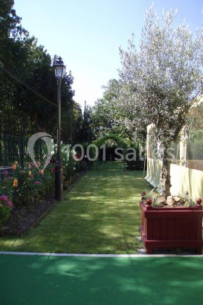 Allée de jardin bordée de fleurs et d'arbres, avec un olivier en pot au premier plan et une arche végétale au fond.