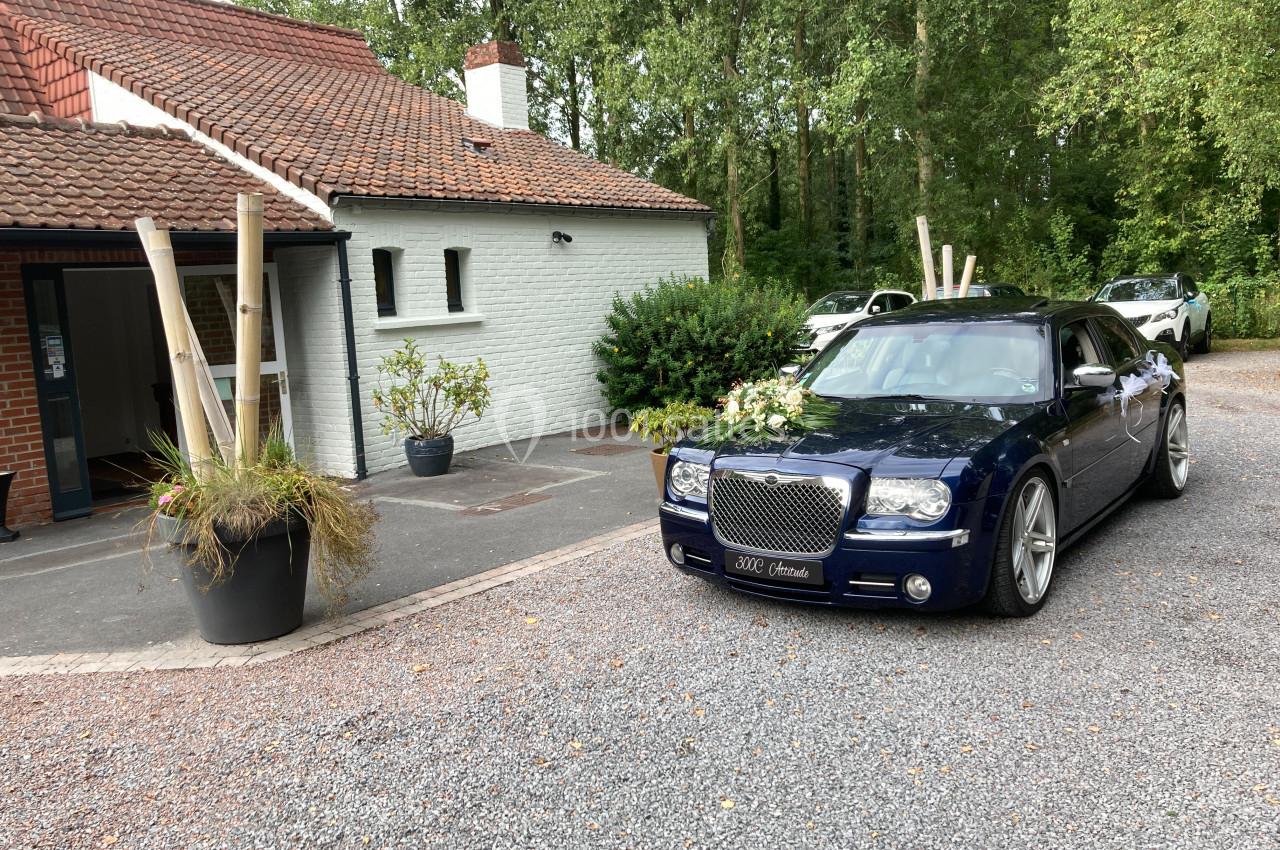 Voiture noire décorée pour un mariage, stationnée devant une maison en briques avec un toit en tuiles rouges.