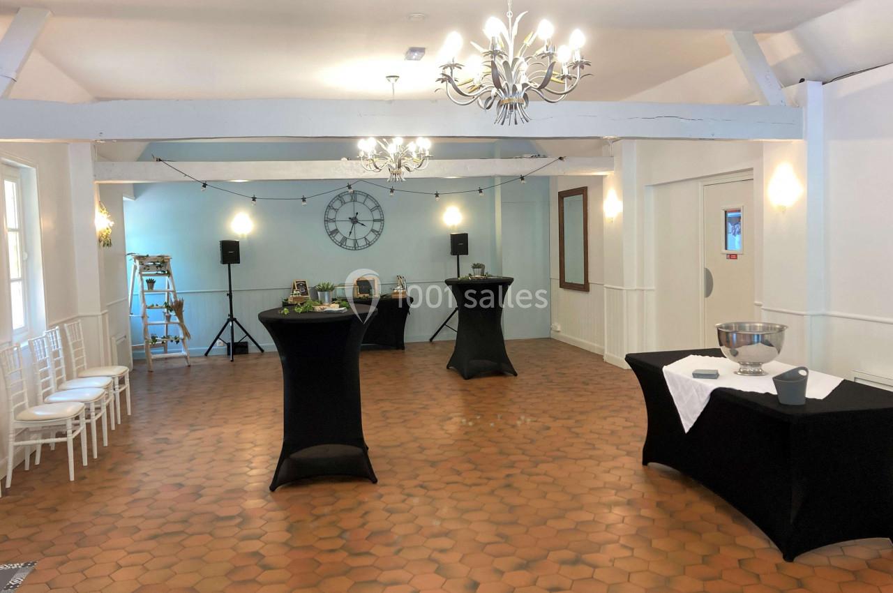 Salle lumineuse avec sol en terre cuite, tables hautes noires, chaises blanches alignées et décoration sobre.