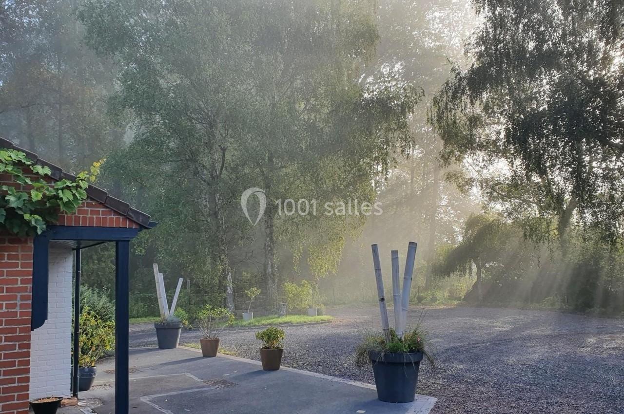 Maison en briques rouges avec terrasse, entourée d'arbres et baignée par des rayons de soleil à travers la brume matinale.