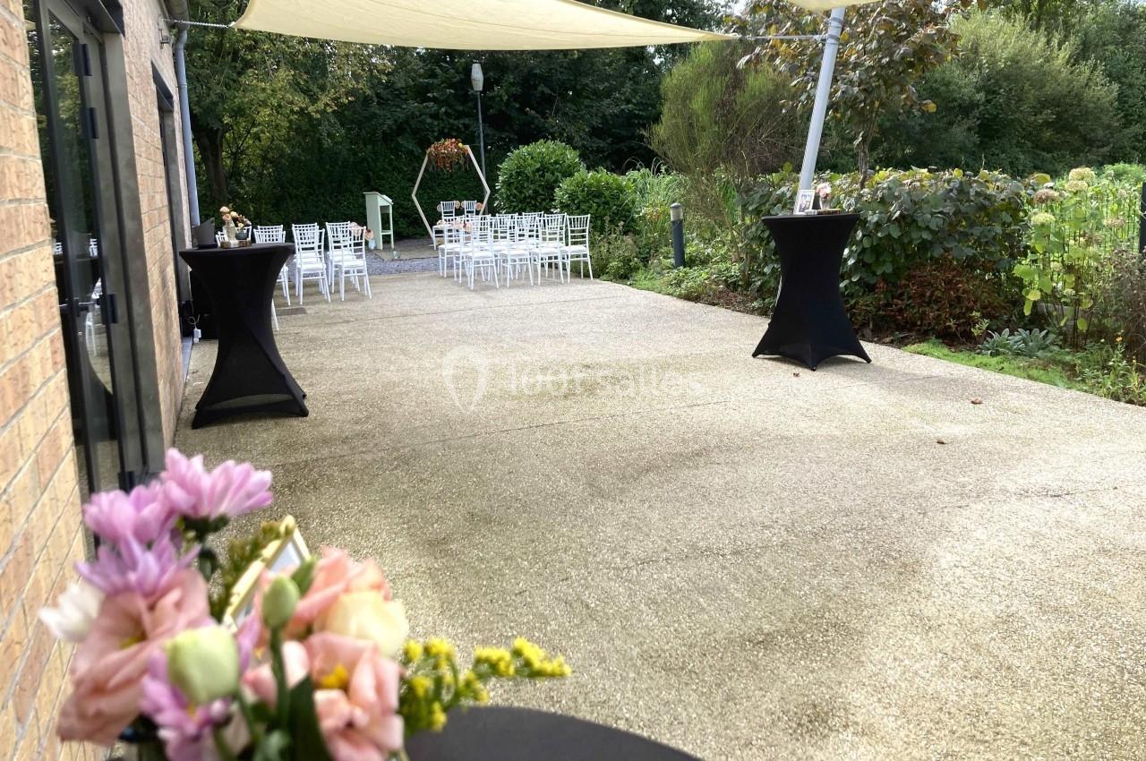 Terrasse aménagée pour un événement avec chaises blanches, tables hautes et décorations florales, entourée de verdure.
