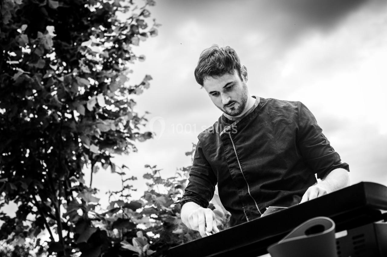 Homme concentré cuisinant en extérieur sur une plancha, entouré de végétation sous un ciel nuageux.