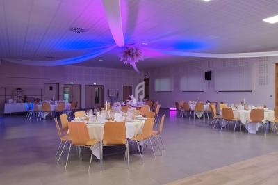 Salle de réception décorée avec des tables rondes dressées, éclairage violet et blanc, plafond orné de voilages.