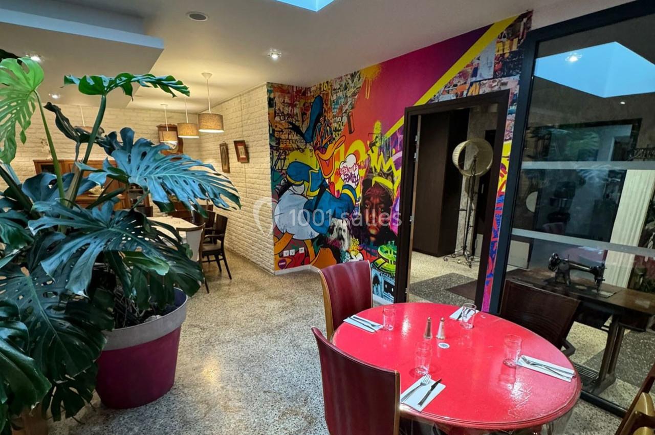 Salle de restaurant colorée avec mur décoré de fresques urbaines, plantes en pot et table rouge dressée.
