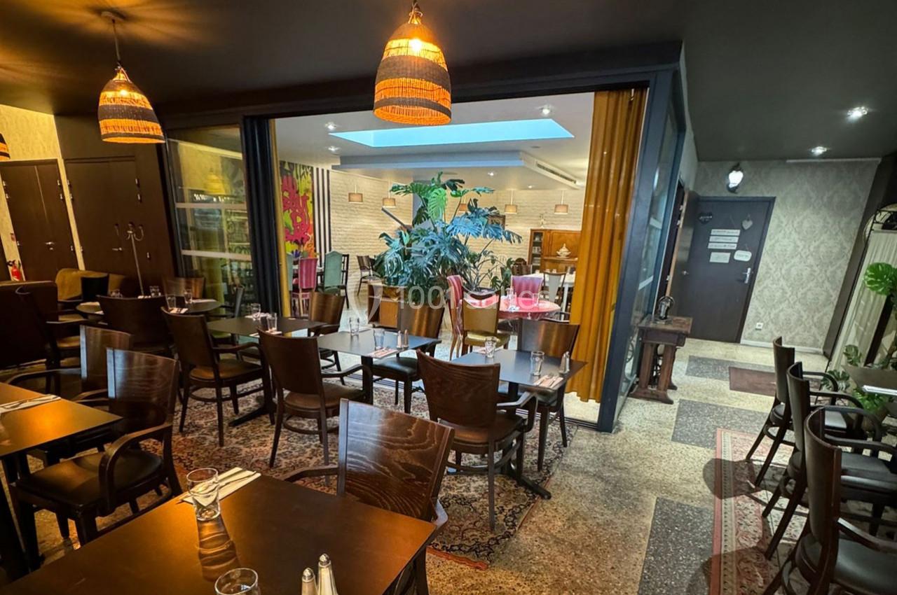 Salle de restaurant chaleureuse avec tables en bois, chaises disposées, plantes et éclairage suspendu.