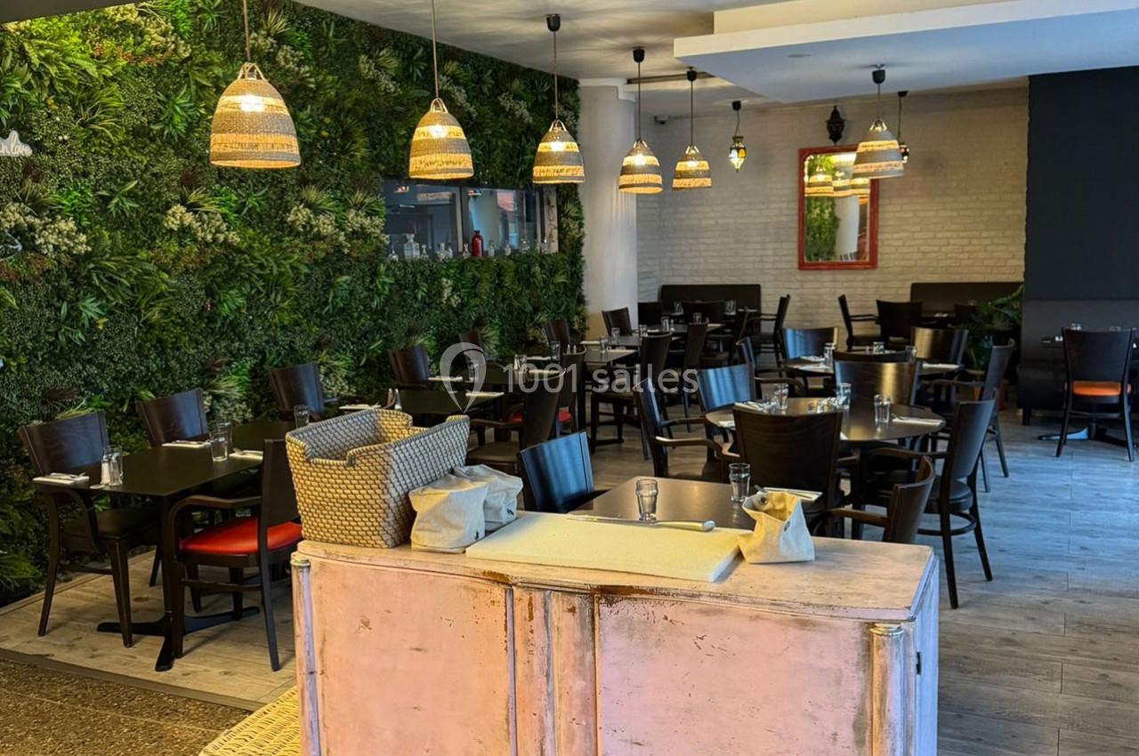 Salle de restaurant avec tables dressées, chaises noires, mur végétal et suspensions lumineuses en osier.