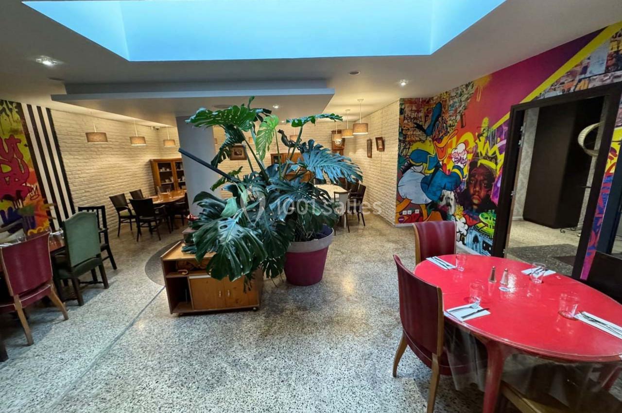 Salle de restaurant avec des tables colorées, une grande plante verte et des murs décorés de fresques artistiques.