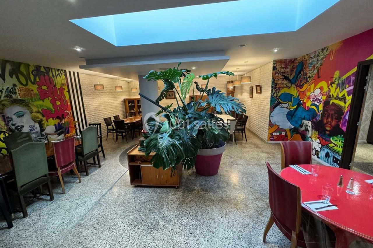 Salle de restaurant colorée avec fresques murales, grandes plantes en pot et tables disposées sous un puits de lumière.