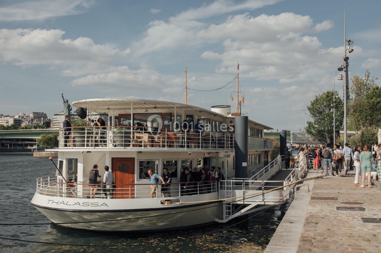 Bateau amarré au bord d'un quai, avec des passagers à bord et des piétons sur la rive sous un ciel dégagé.