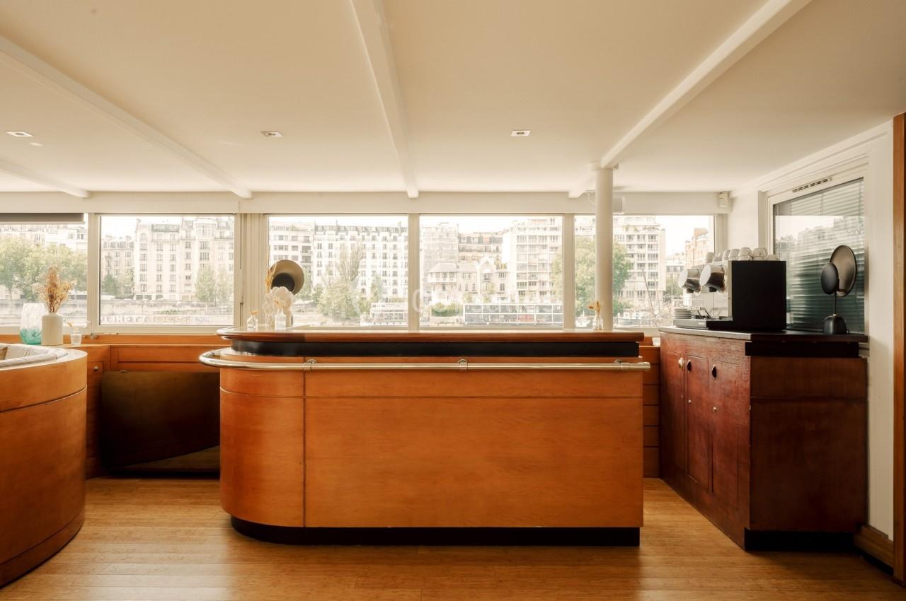 Intérieur lumineux d'un bar en bois avec vue sur des bâtiments et la Seine à travers de grandes baies vitrées.