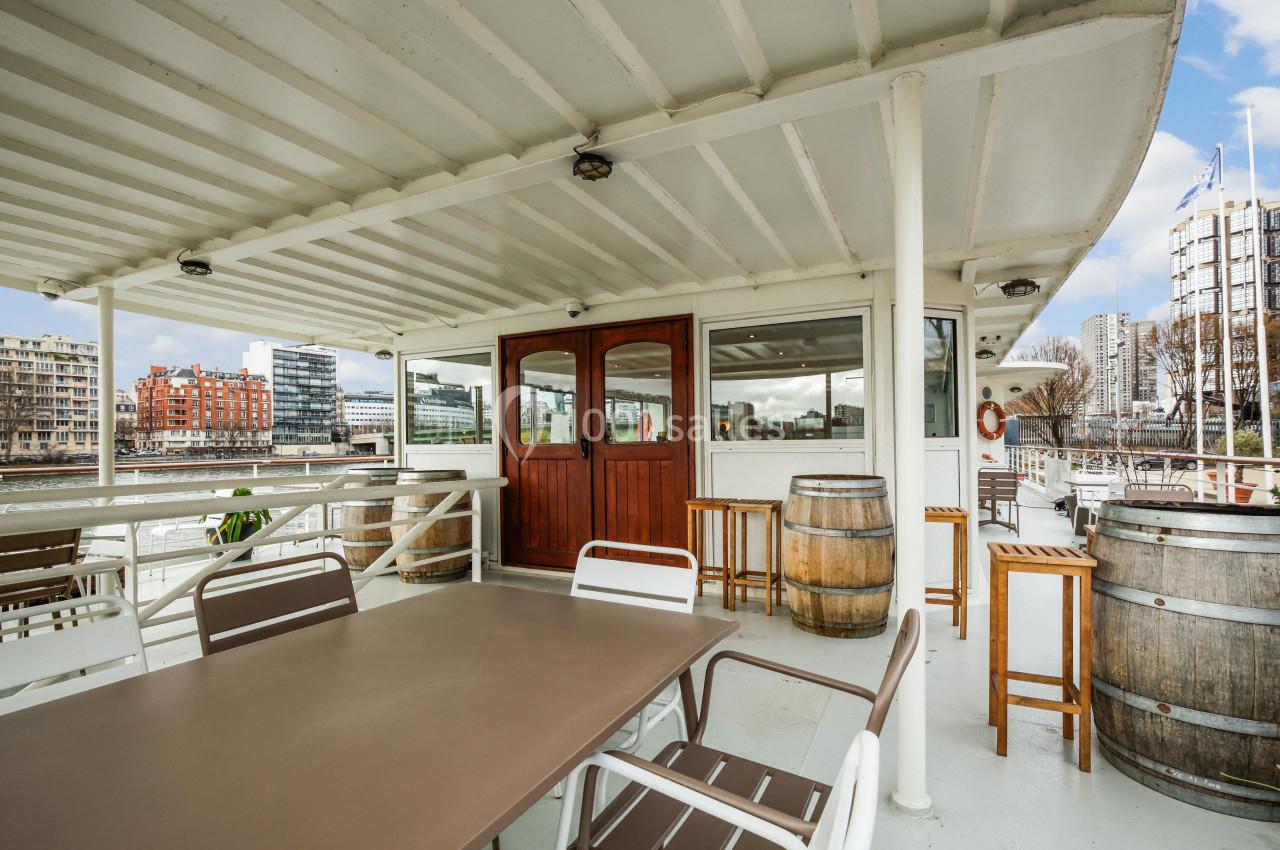 Terrasse d'un bateau avec tables, chaises, tonneaux en bois et vue sur des immeubles en bord de rivière.