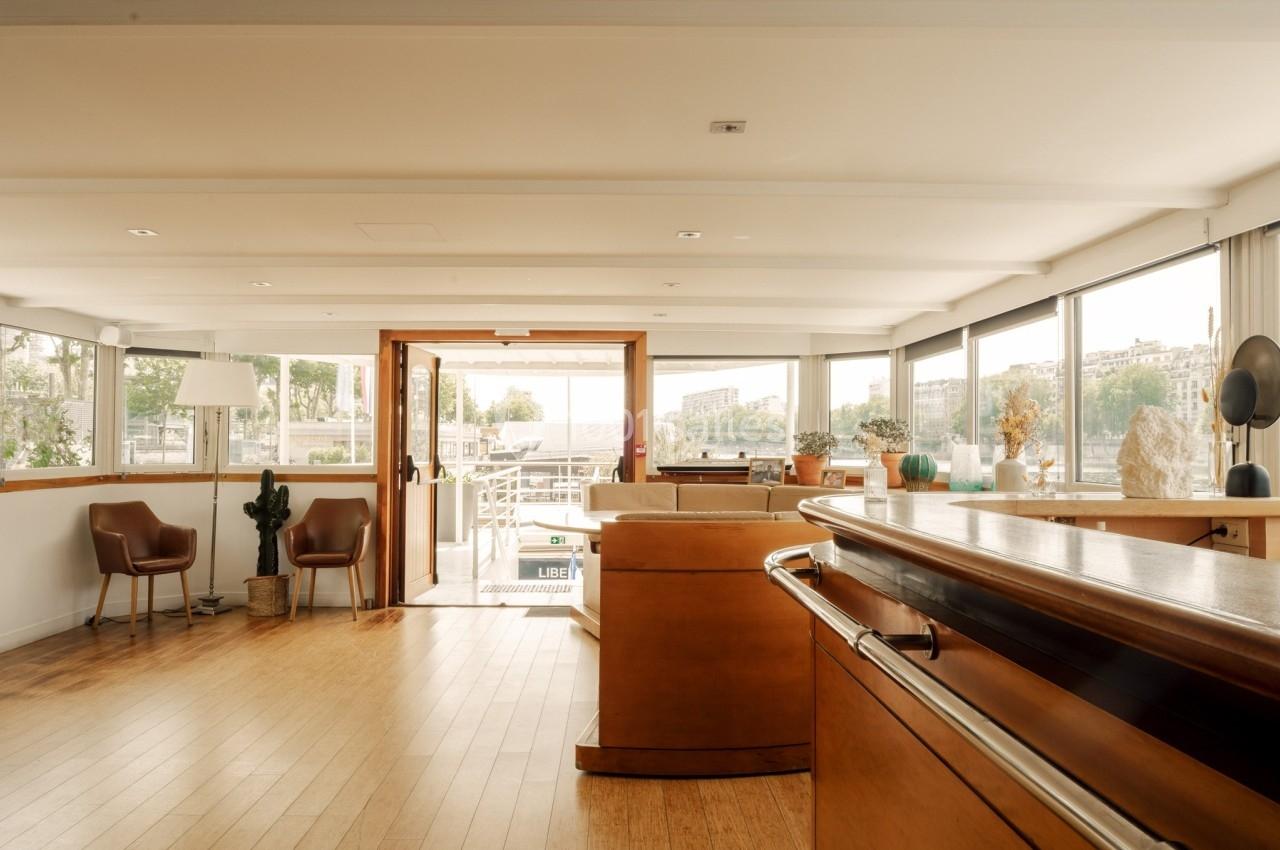 Intérieur lumineux d'une péniche avec parquet, mobilier en bois et grandes baies vitrées donnant sur l'extérieur.