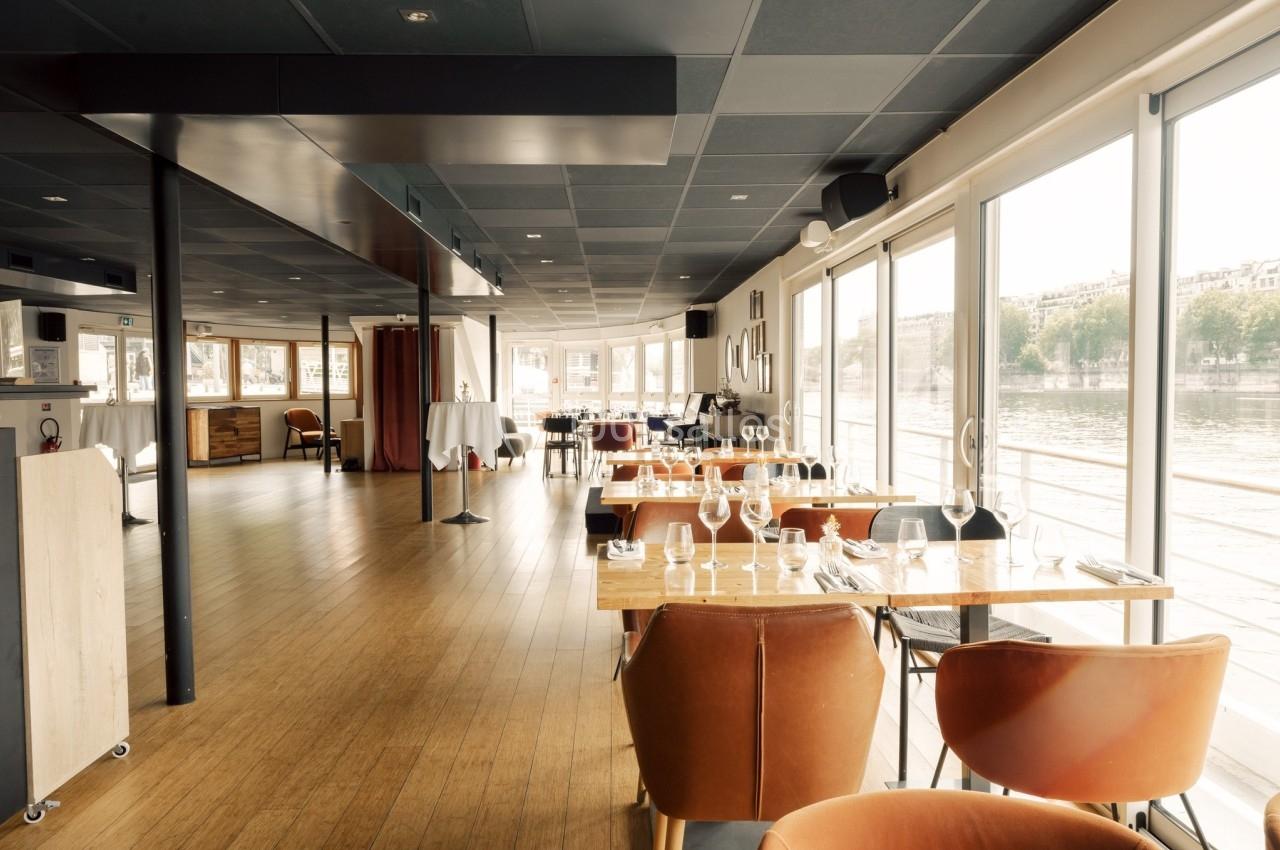 Salle de restaurant lumineuse avec grandes baies vitrées donnant sur une vue extérieure fluviale.