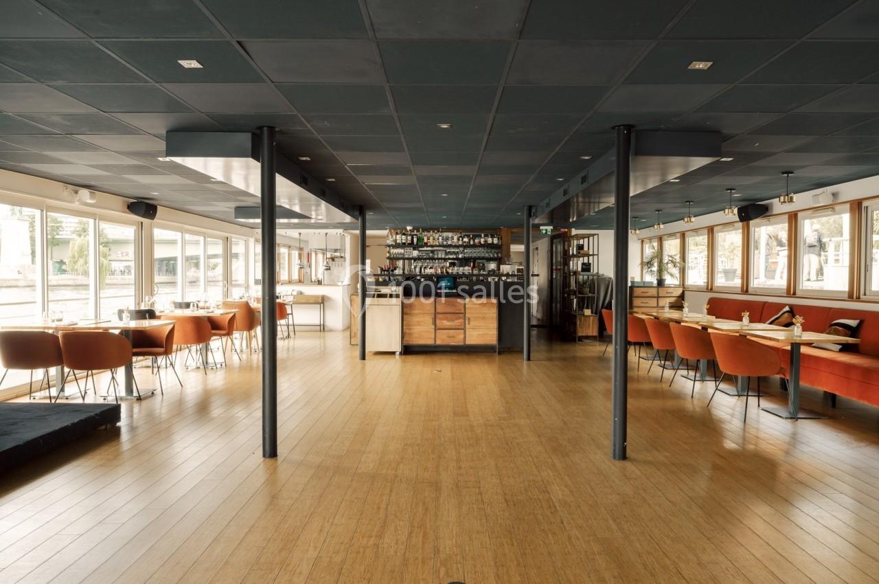 Salle de restaurant lumineuse avec parquet en bois, tables alignées et un bar au fond.