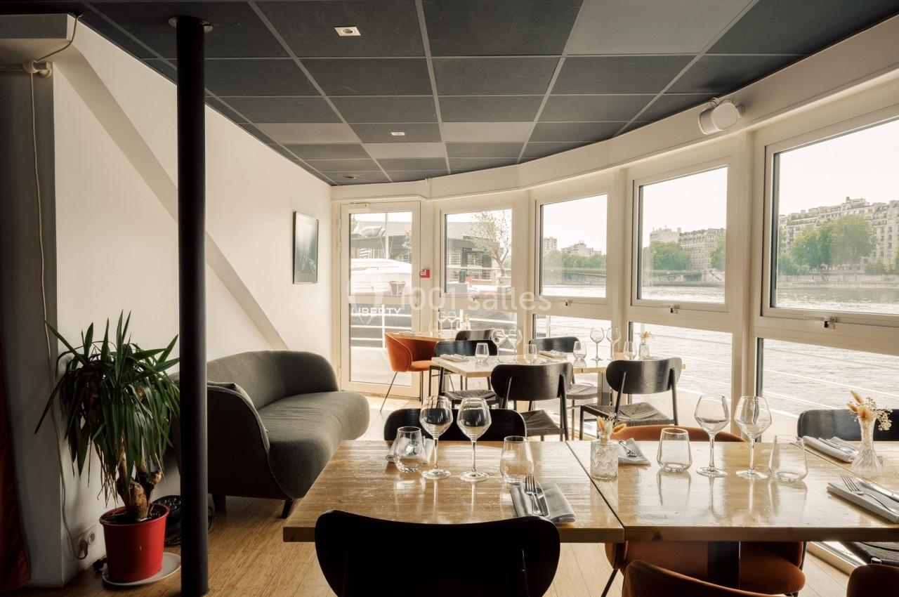 Salle de restaurant lumineuse avec grandes baies vitrées, tables dressées et vue sur un fleuve en arrière-plan.