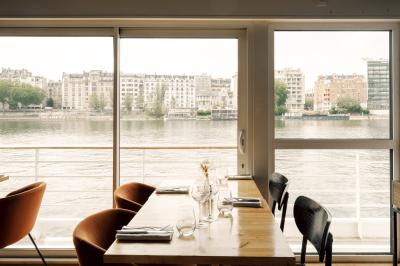 Salle de restaurant lumineuse avec des tables dressées, des chaises modernes et vue sur une rivière bordée de bâtiments.