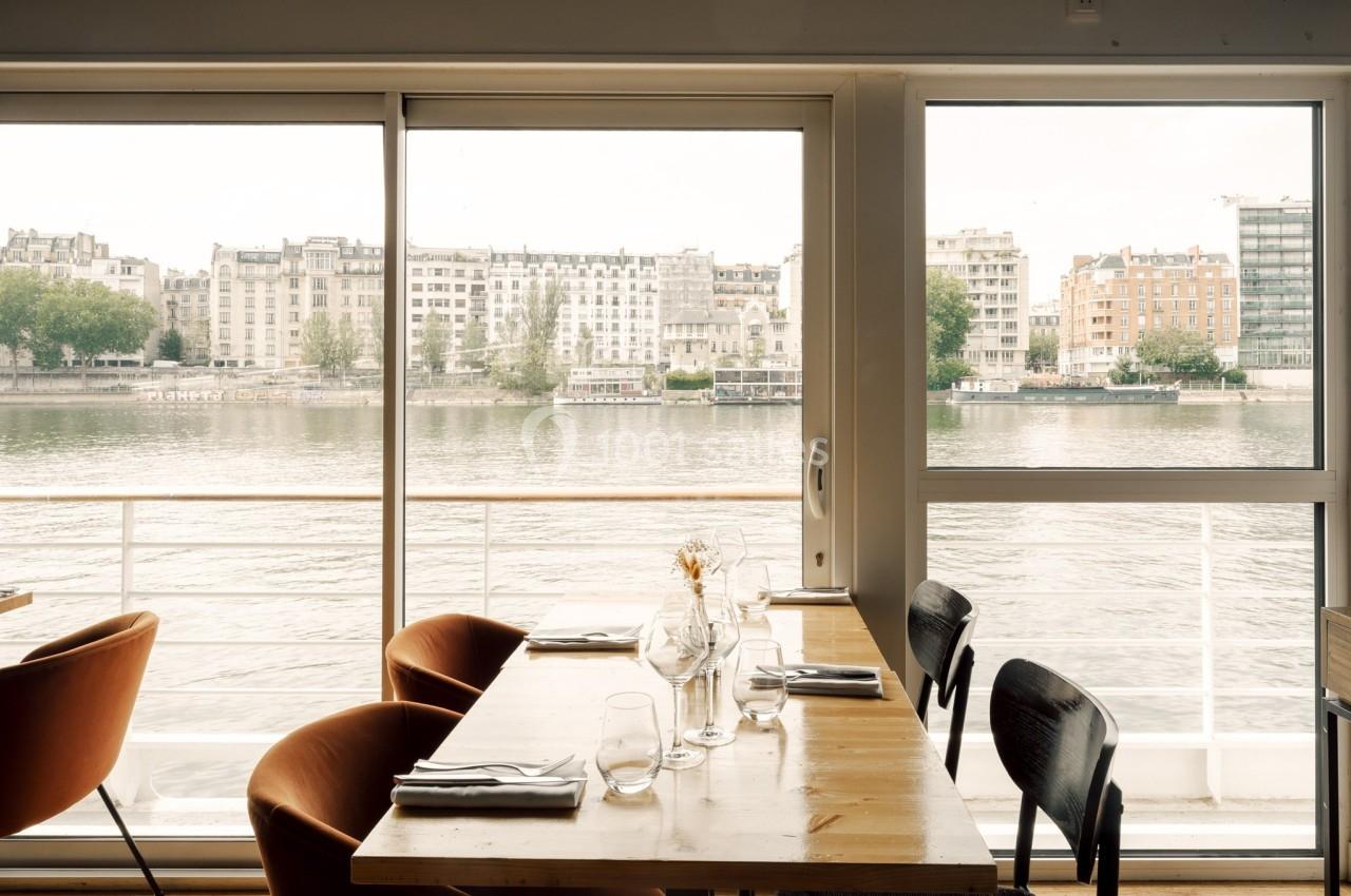 Salle de restaurant avec vue sur une rivière et des immeubles en arrière-plan, à travers de grandes baies vitrées.