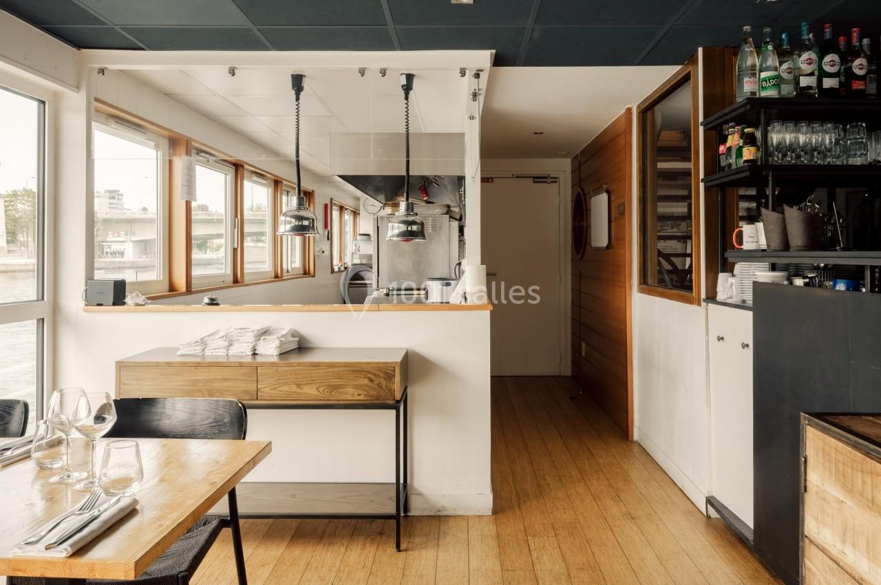 Salle de restaurant lumineuse avec table dressée, cuisine ouverte en arrière-plan et étagères avec verres et bouteilles.