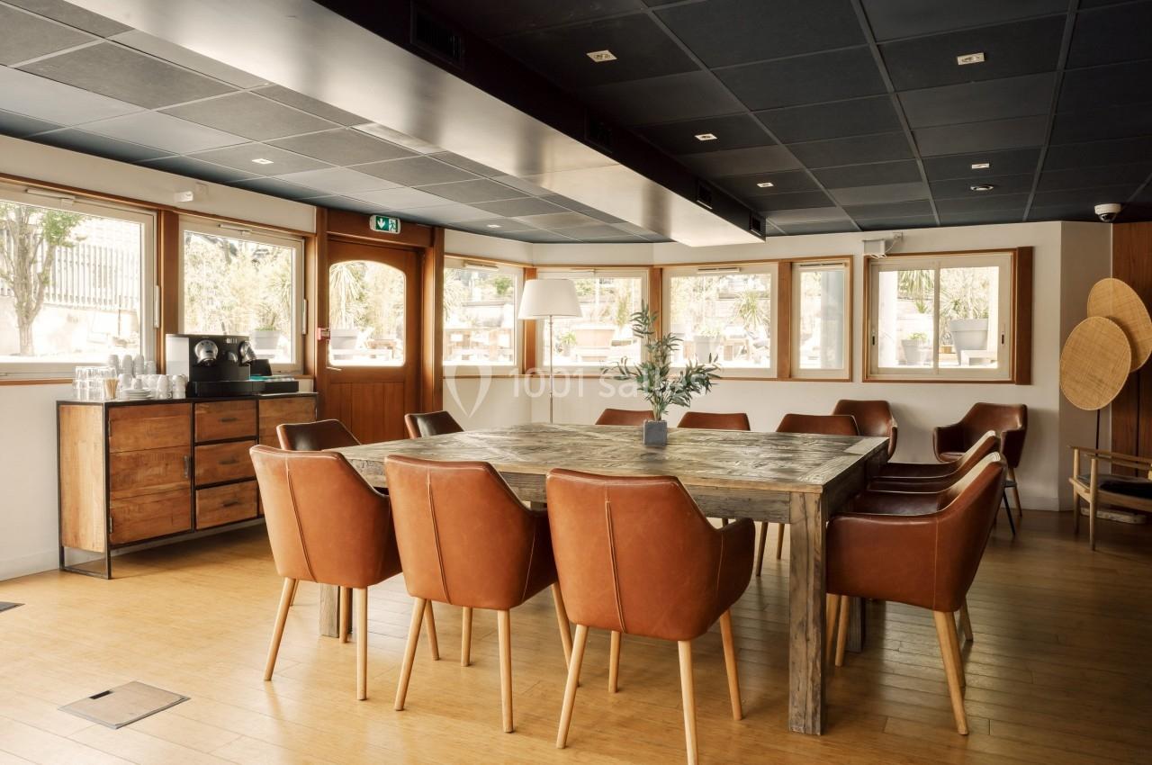 Salle lumineuse avec une grande table en bois entourée de chaises en cuir marron, décorée d'une plante au centre.