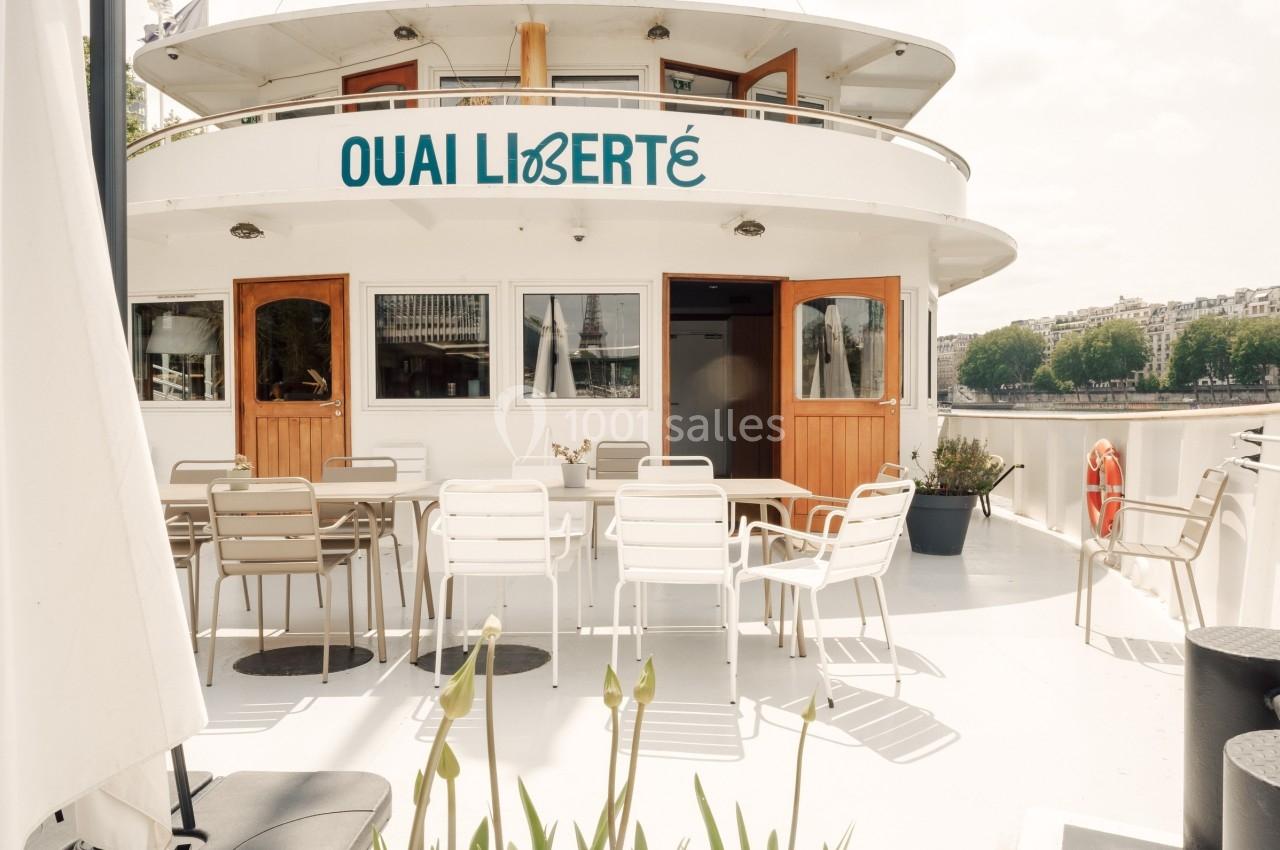 Terrasse d'un bateau aménagée avec des tables et chaises, amarré au bord d'une rivière en milieu urbain.