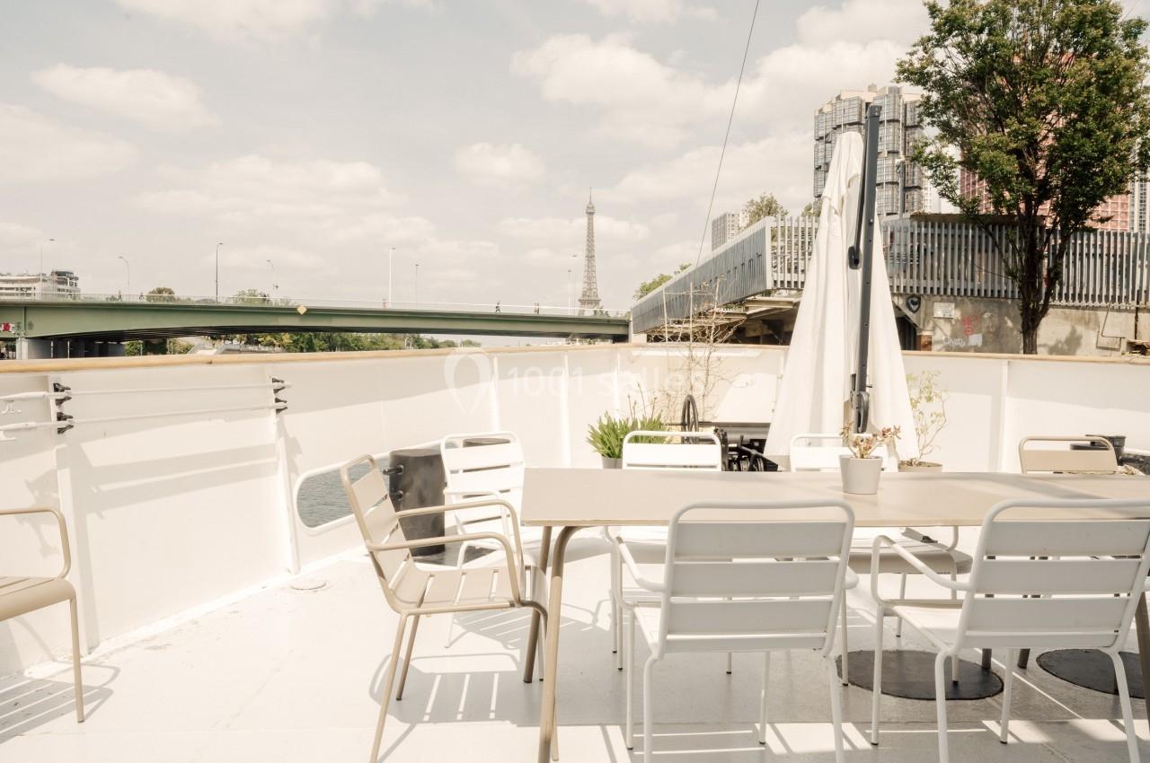 Terrasse extérieure d'un bateau avec table, chaises et parasol, vue sur un pont et la tour Eiffel en arrière-plan.