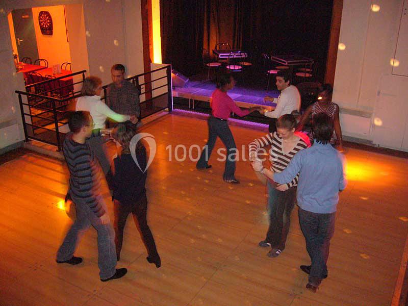 Des personnes dansent en couple sur une piste en bois éclairée, avec des tables et une scène en arrière-plan.