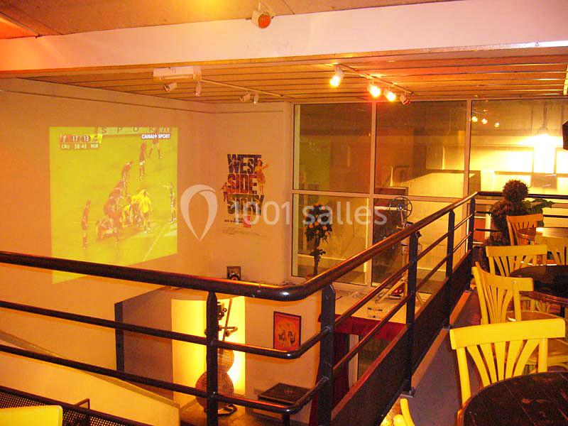 Salle avec des tables et chaises, écran projetant un match de rugby, éclairage chaleureux et décoration moderne.