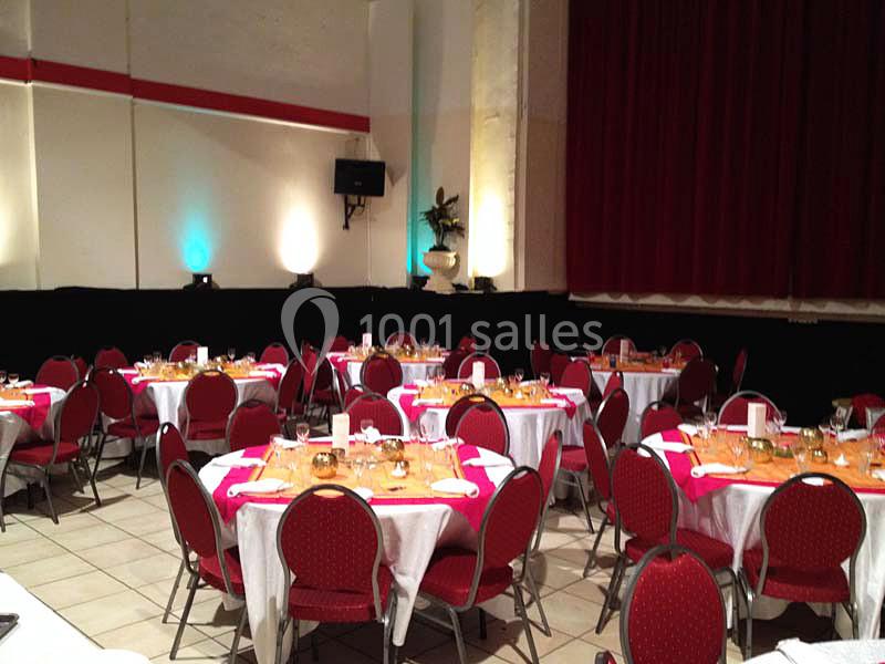 Salle de réception avec des tables rondes dressées, nappes blanches et roses, chaises rouges et éclairage tamisé.