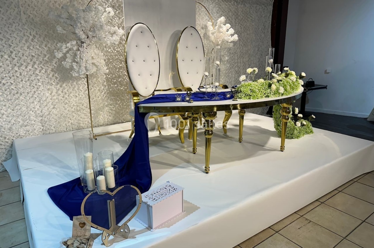 Table décorée pour un mariage avec chaises blanches, nappe bleue, fleurs blanches et éléments dorés sur une estrade.