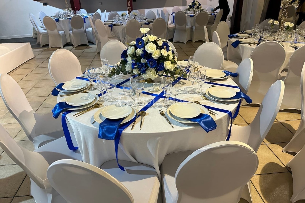 Table ronde décorée pour un événement, avec nappes blanches, rubans bleus, vaisselle élégante et centre de table floral.