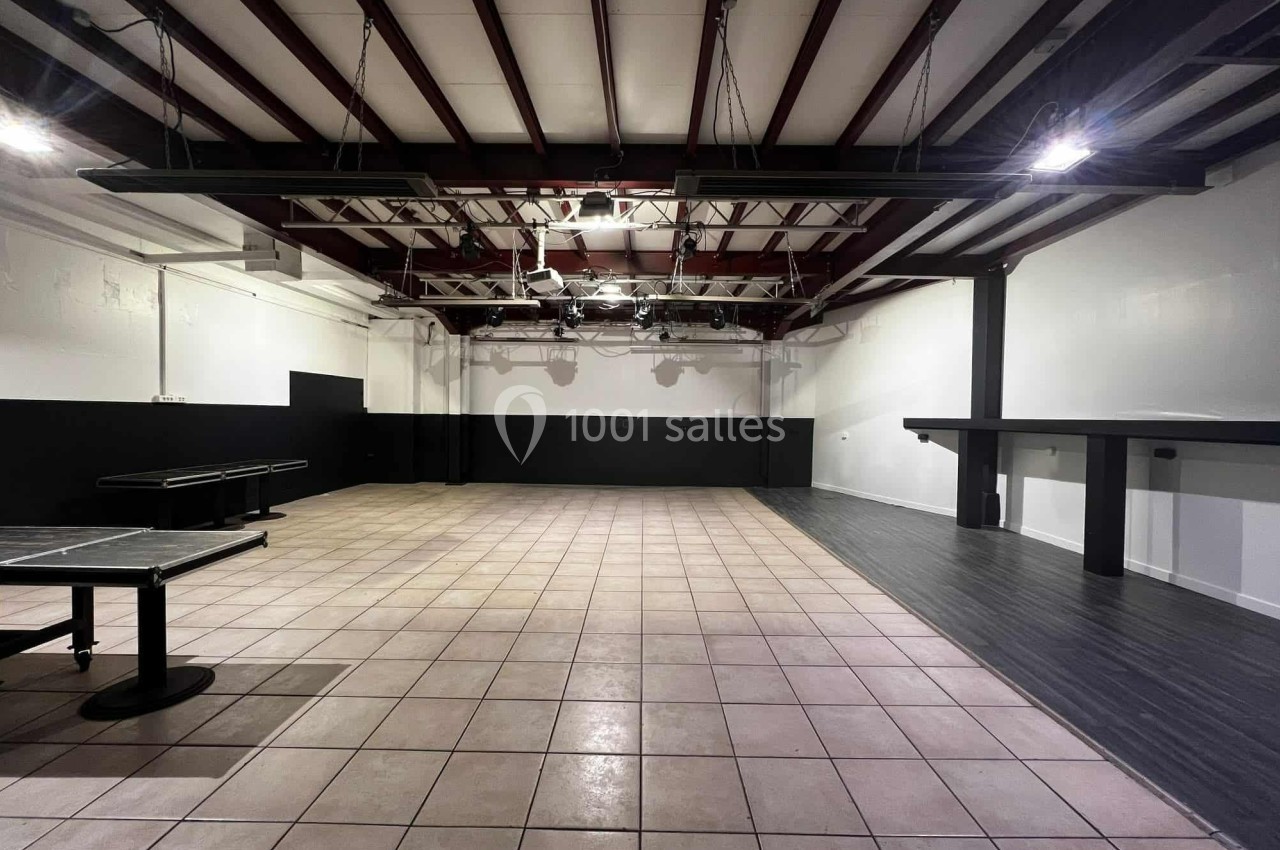 Salle vide avec sol en carrelage et parquet, murs blancs et noirs, plafond avec poutres apparentes et éclairage suspendu.