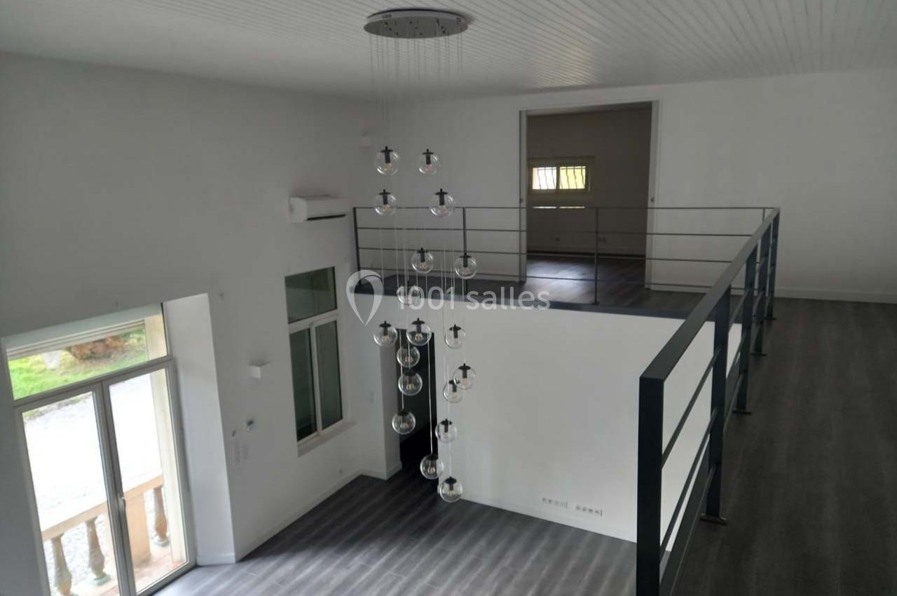 Intérieur d'une pièce lumineuse avec mezzanine, sol en parquet gris, grandes fenêtres et suspension moderne.