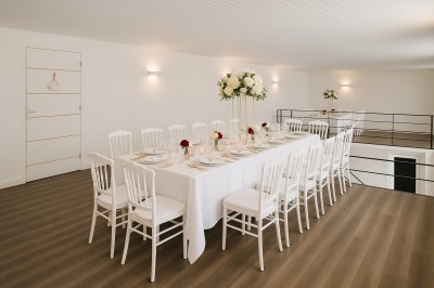 Salle lumineuse avec une grande table dressée pour un repas, décorée de fleurs blanches et rouges, entourée de chaises…