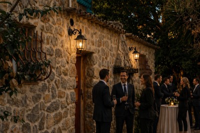 Personnes en tenue formelle discutant devant une maison en pierre éclairée, lors d'un événement en extérieur.