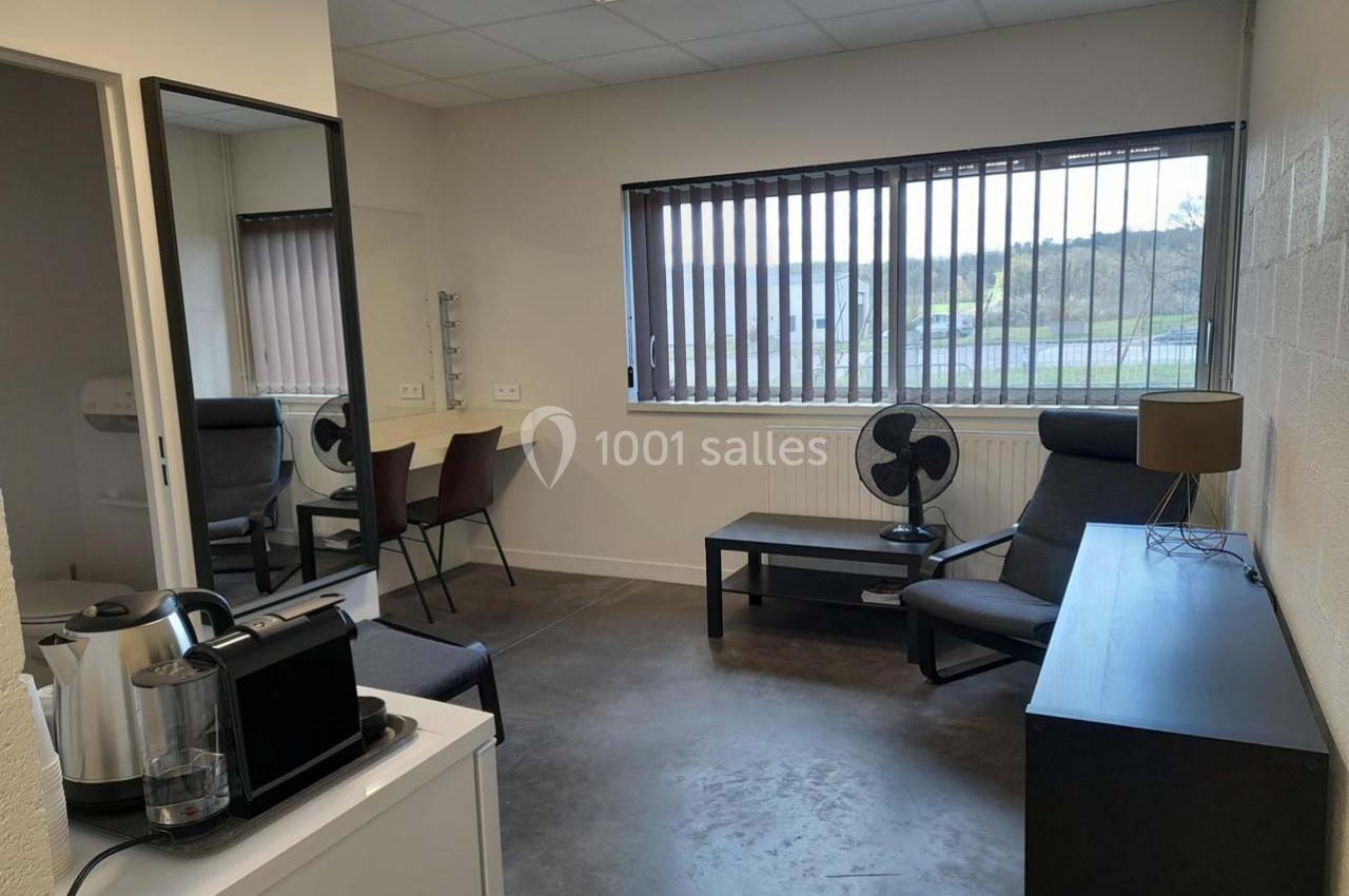 Salle lumineuse avec bureau, fauteuil, miroir, cafetière et fenêtre à stores donnant sur un extérieur verdoyant.