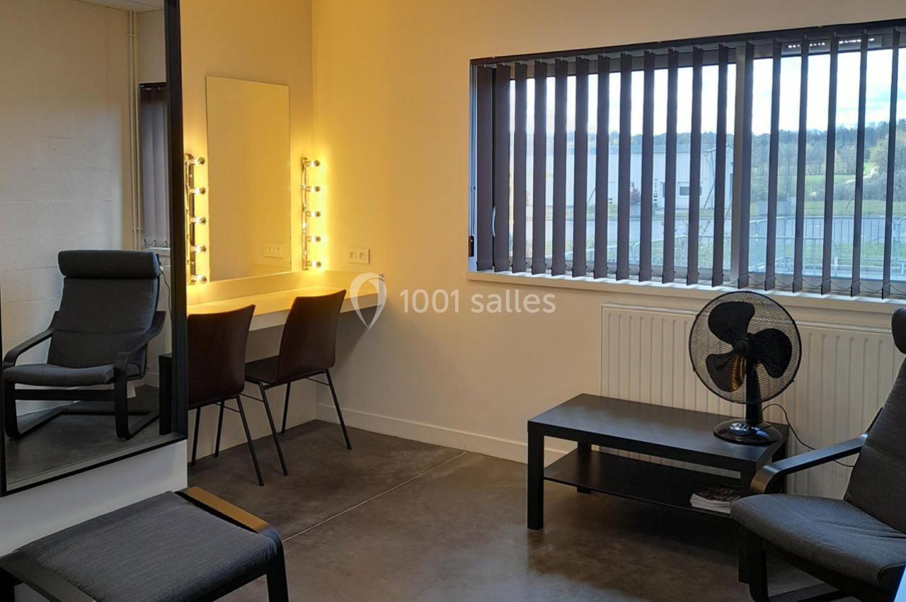 Salle de loge avec miroir éclairé, chaises, table basse, ventilateur et fenêtre avec stores donnant sur l'extérieur.