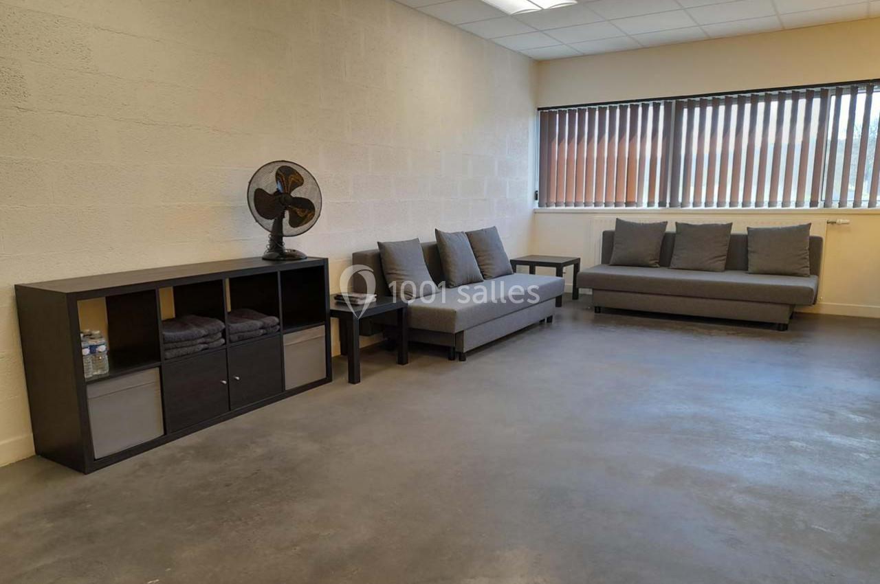 Salle lumineuse avec deux canapés gris, une étagère noire, un ventilateur et un sol en béton poli.