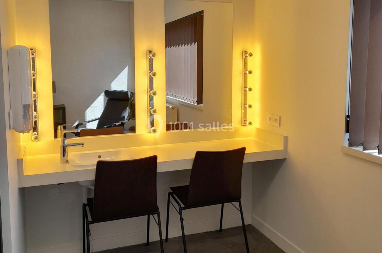 Coiffeuse avec deux chaises, miroir éclairé par des ampoules et lavabo intégré, dans une pièce lumineuse.