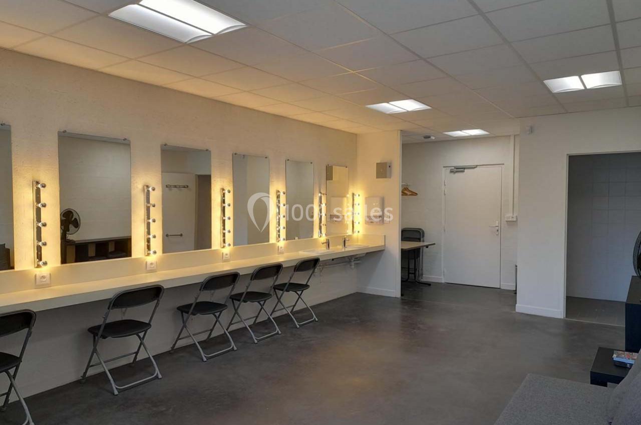 Salle de loges avec miroirs éclairés, chaises pliantes et éclairage au plafond, ambiance sobre et fonctionnelle.