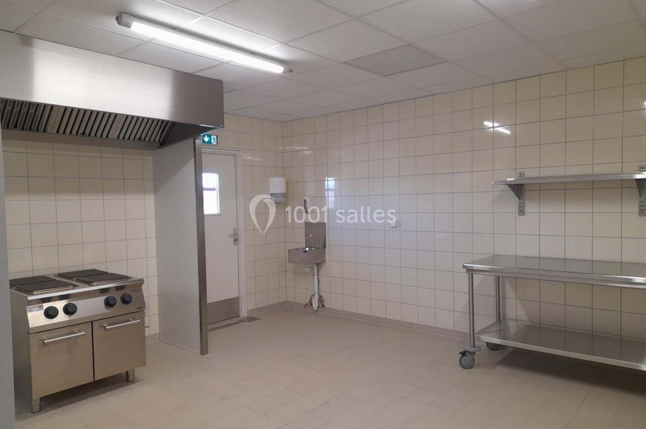 Cuisine professionnelle vide avec plaques de cuisson, évier, étagère en inox et murs carrelés blancs.