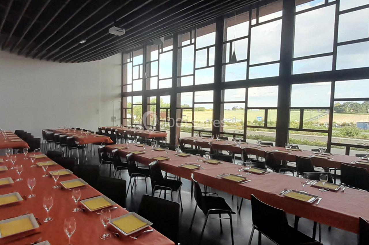 Salle de réception lumineuse avec de grandes baies vitrées, tables dressées avec nappes rouges, verres et couverts.