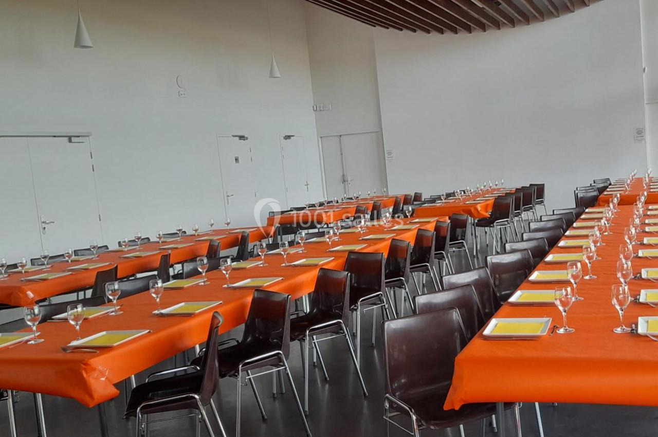 Salle aménagée avec des tables rectangulaires couvertes de nappes orange, disposées en rangées, avec chaises et verres.