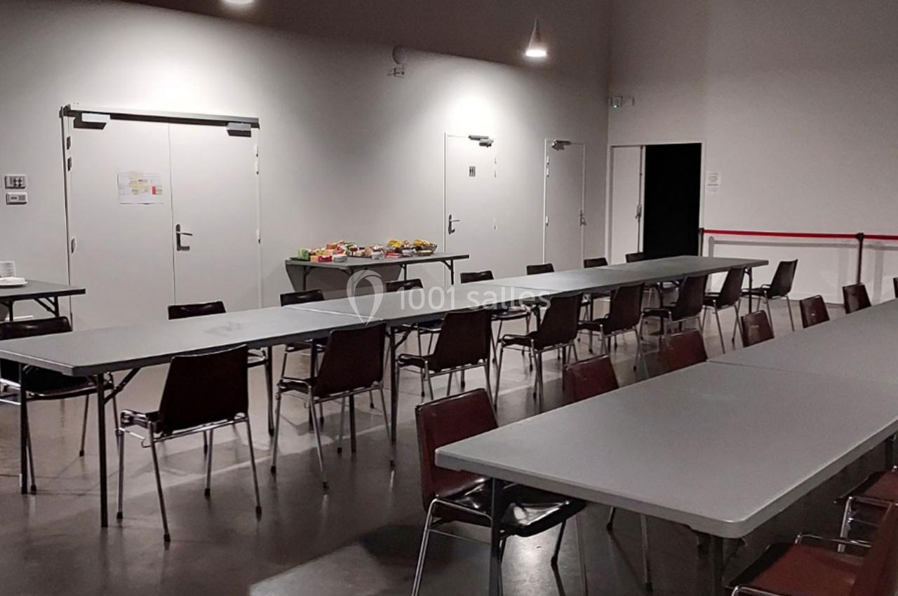 Salle avec de longues tables et des chaises alignées, un buffet disposé sur une table au fond près des portes.