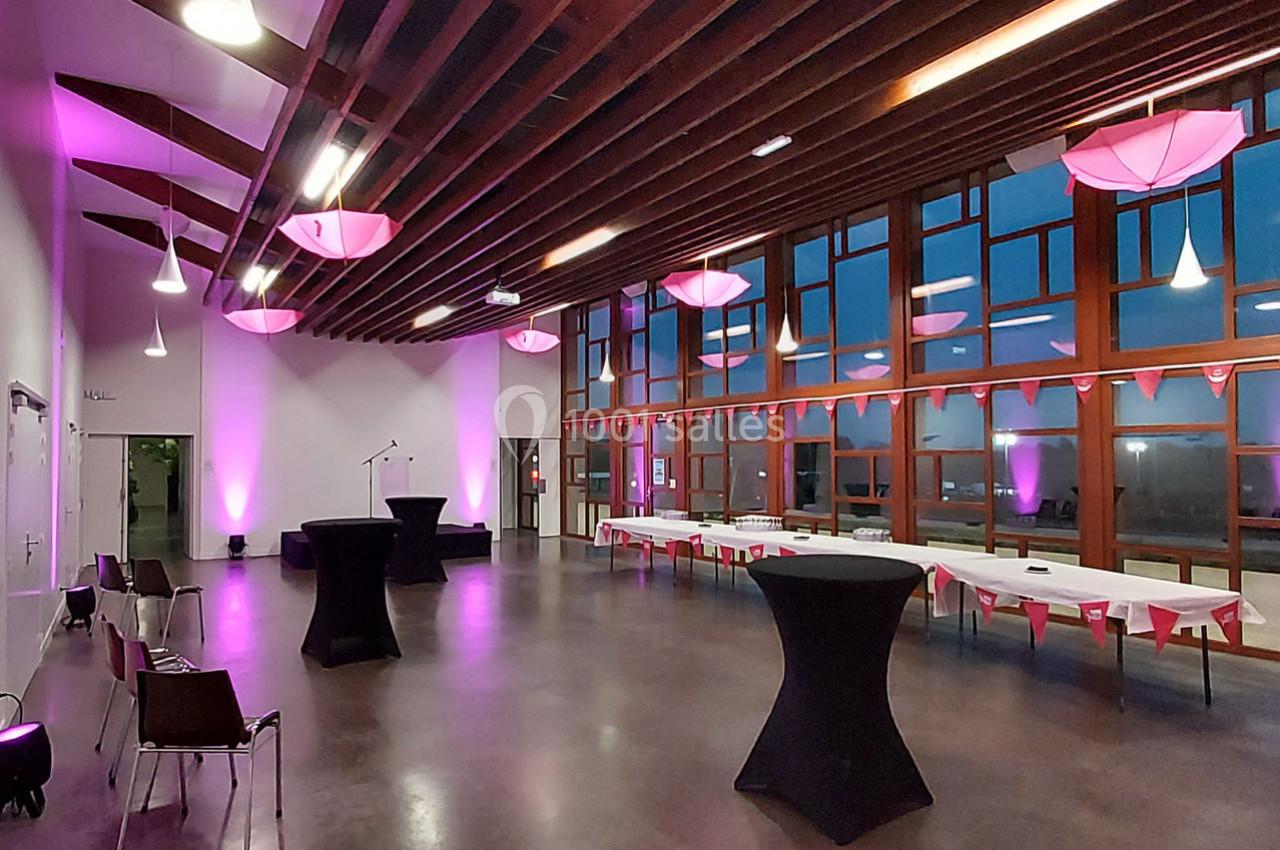 Salle de réception moderne avec éclairage rose, grandes baies vitrées et tables décorées pour un événement.