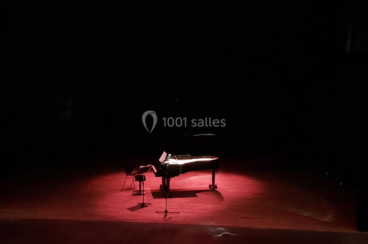 Un piano à queue éclairé par une lumière rouge sur une scène sombre, avec un tabouret et un pupitre.