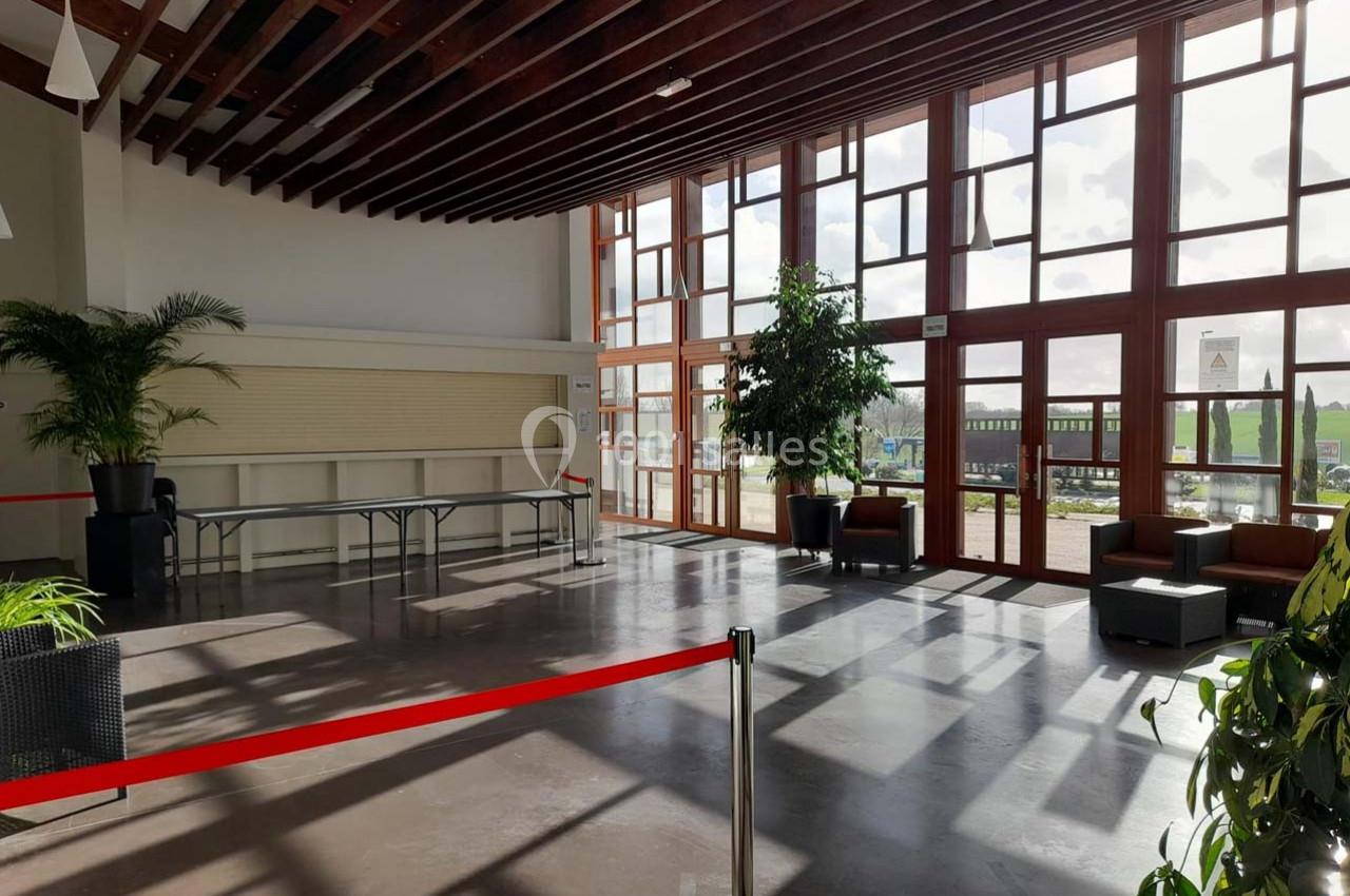 Salle lumineuse avec de grandes baies vitrées, des plantes, des fauteuils et une table pliante près d'un mur.